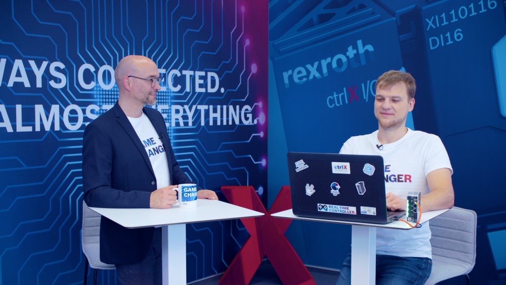Gastgeber Christian Zentraf und sein Gast Michael Schäfer stehen an Besprechungstischen und sprechen über das Programmieren von Python-Skripten direkt auf der ctrlX CORE, der Steuerungshardware der Automatisierungslösung ctrlX AUTOMATION