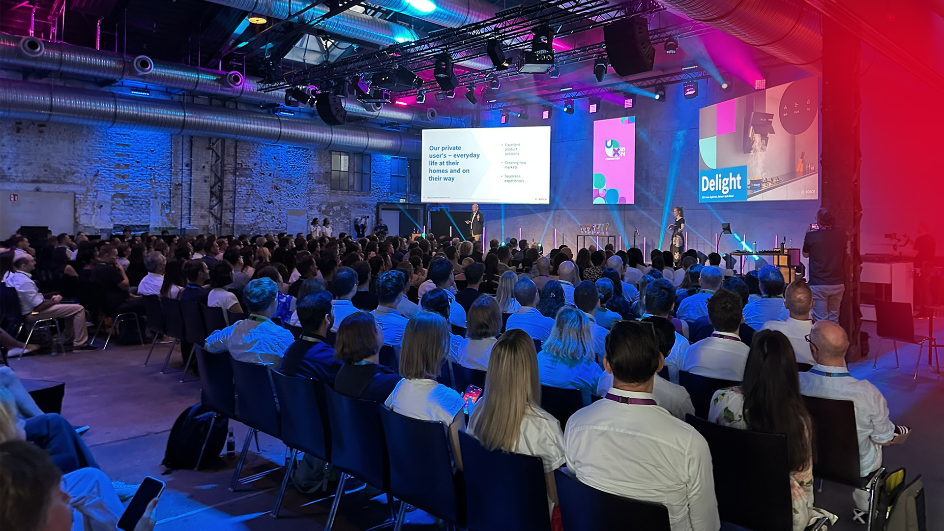 Das Bild zeigt die Räumlichkeiten Bosch UX Convention. Zwei Redner:innen präsentieren auf der Bühne vor den Teilnehmer:innen.